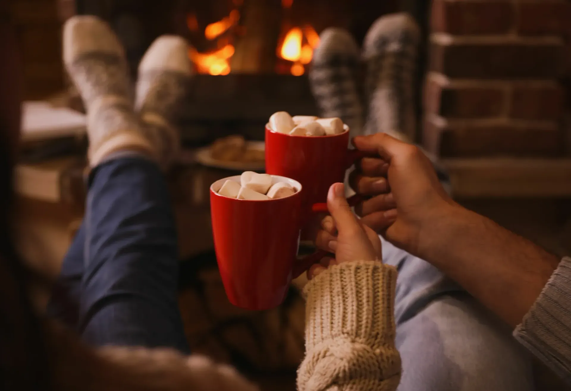 A couple is sitting in front of a fireplace drinking hot chocolate with marshmallows.