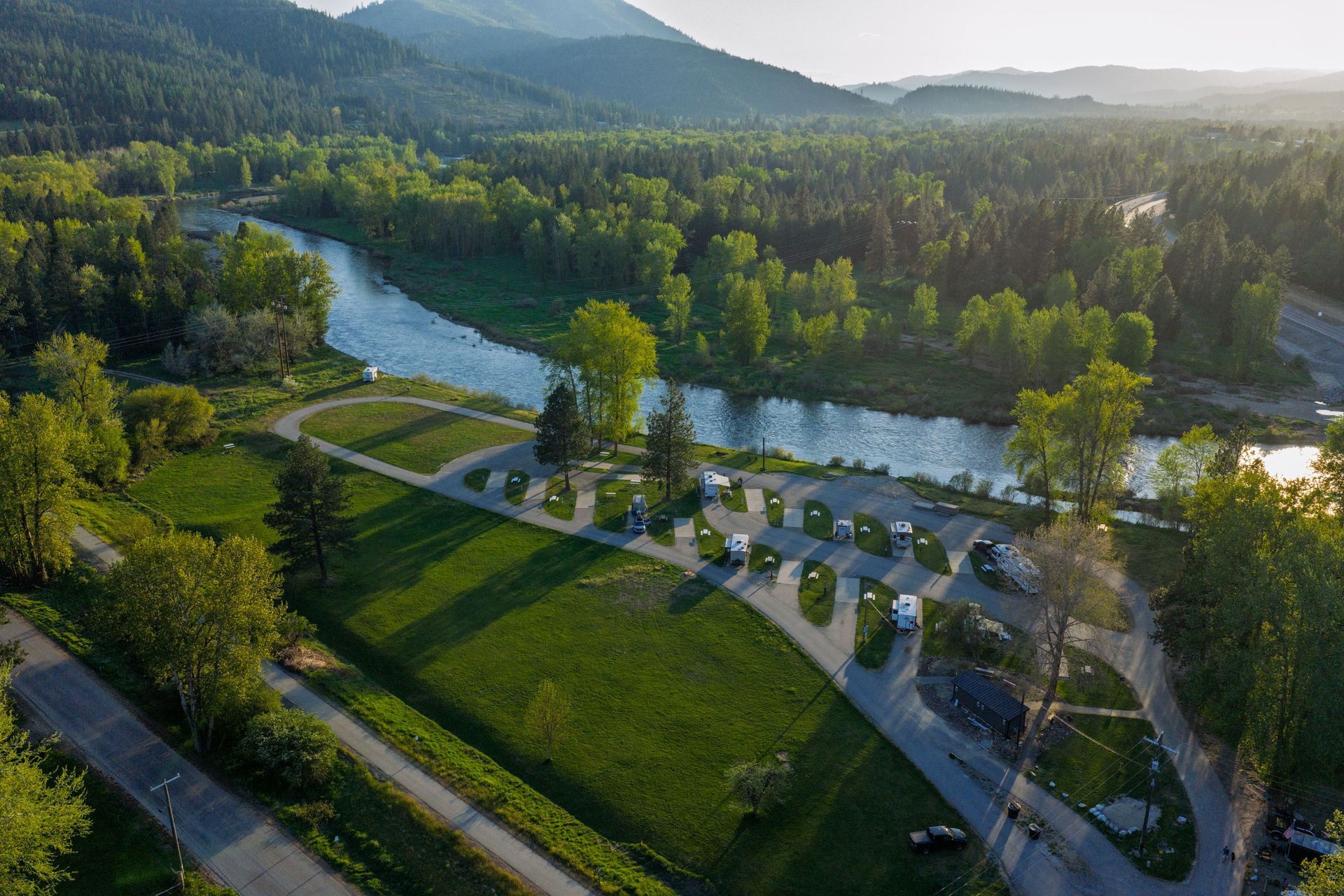 Aerial view of a green riverside campground with winding roads, tents, and forested hills in the background