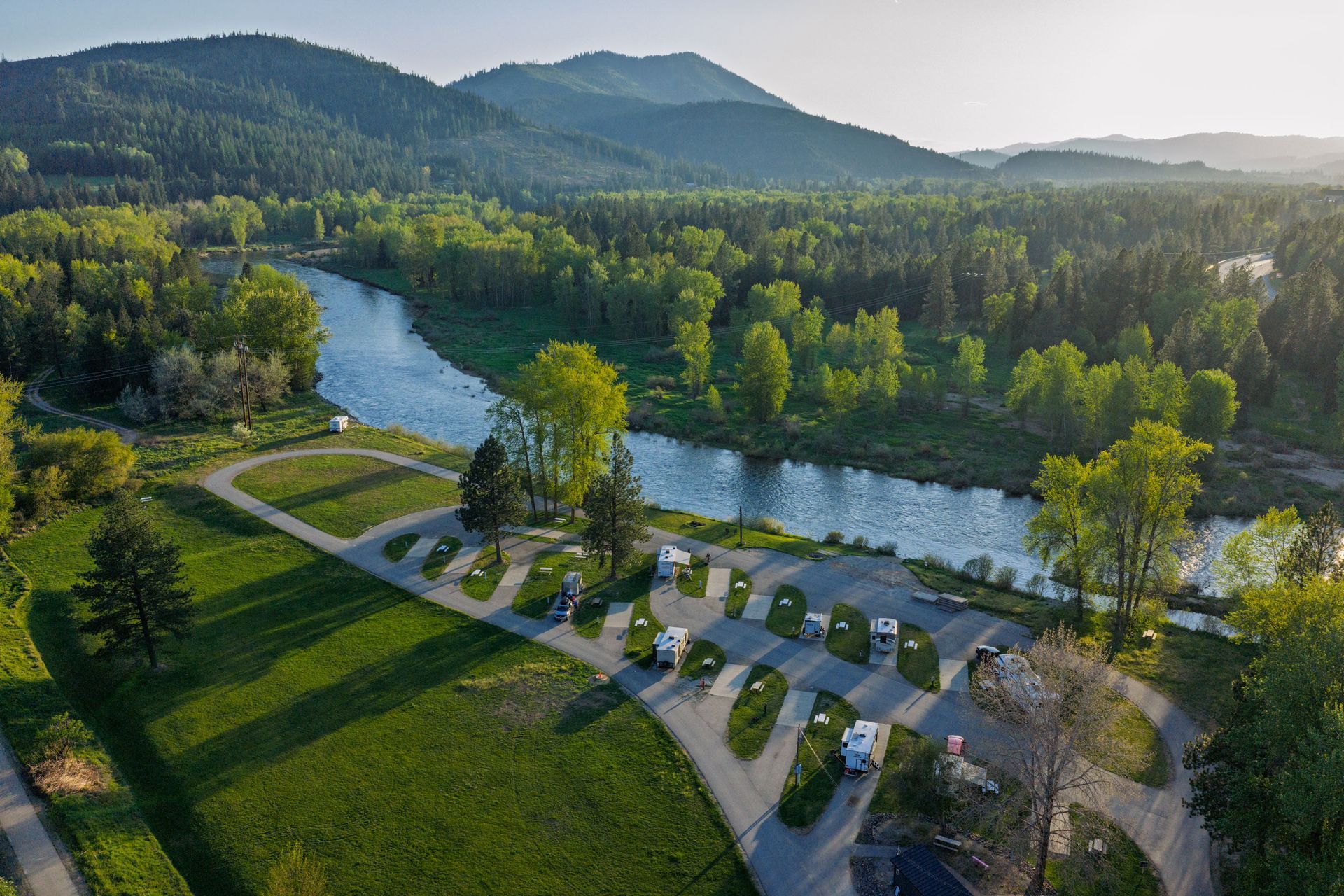 Aerial view of a river, parking area, and green park with hills in the background.