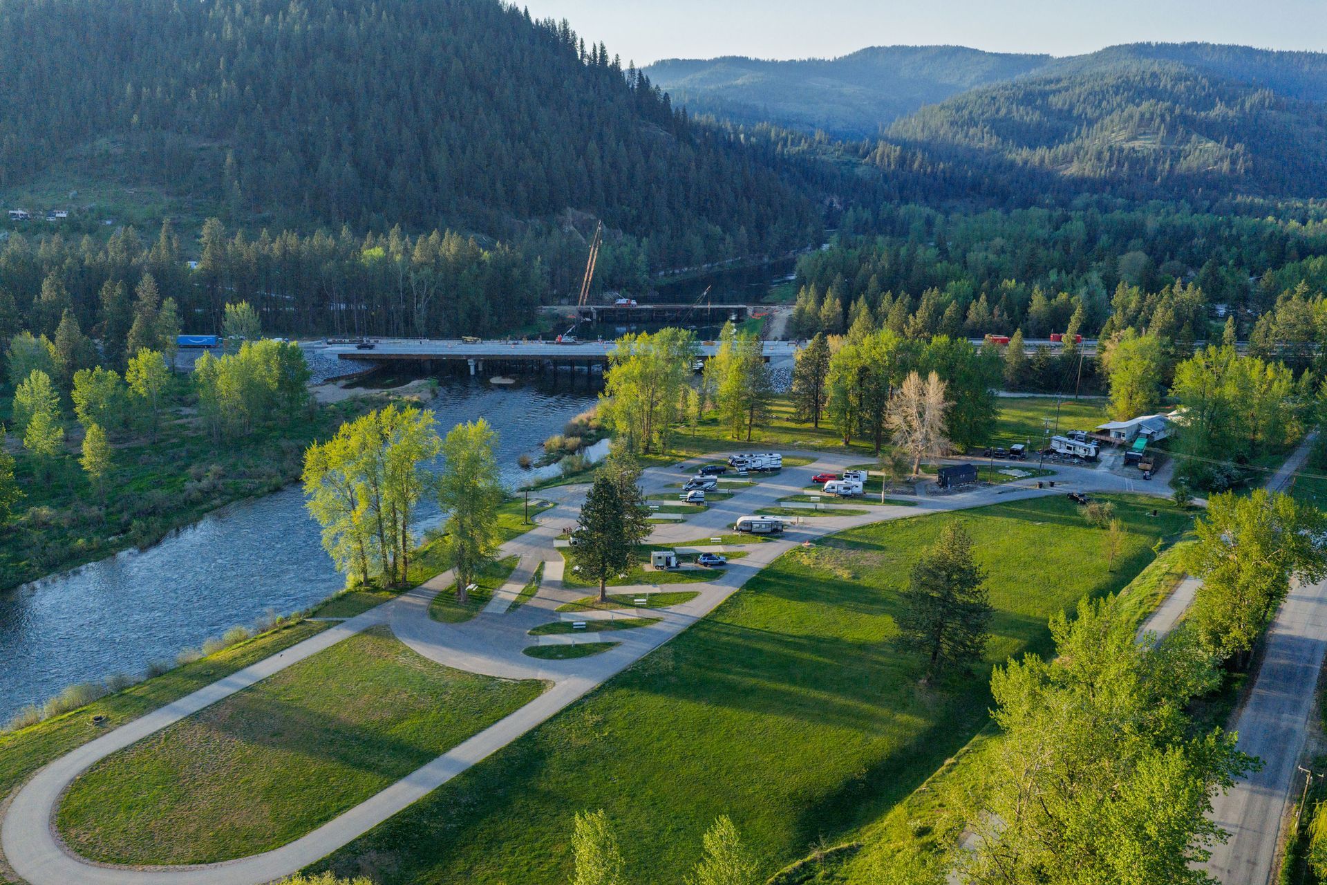 Aerial view of a riverside park with paths, trees, and a bridge, backed by forested hills.