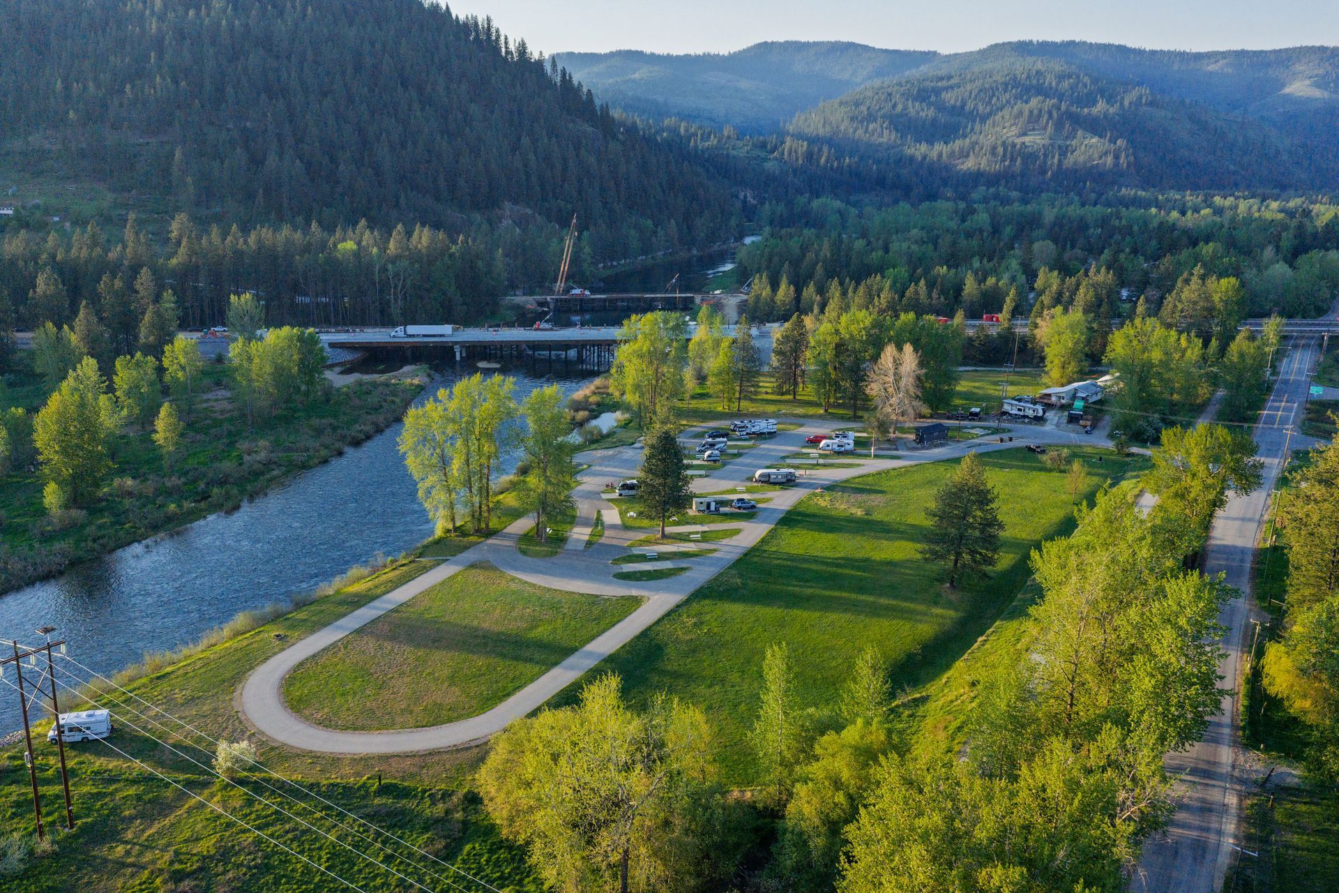 Aerial view of a riverside park with winding paths, trees, a road, and forested hills beyond