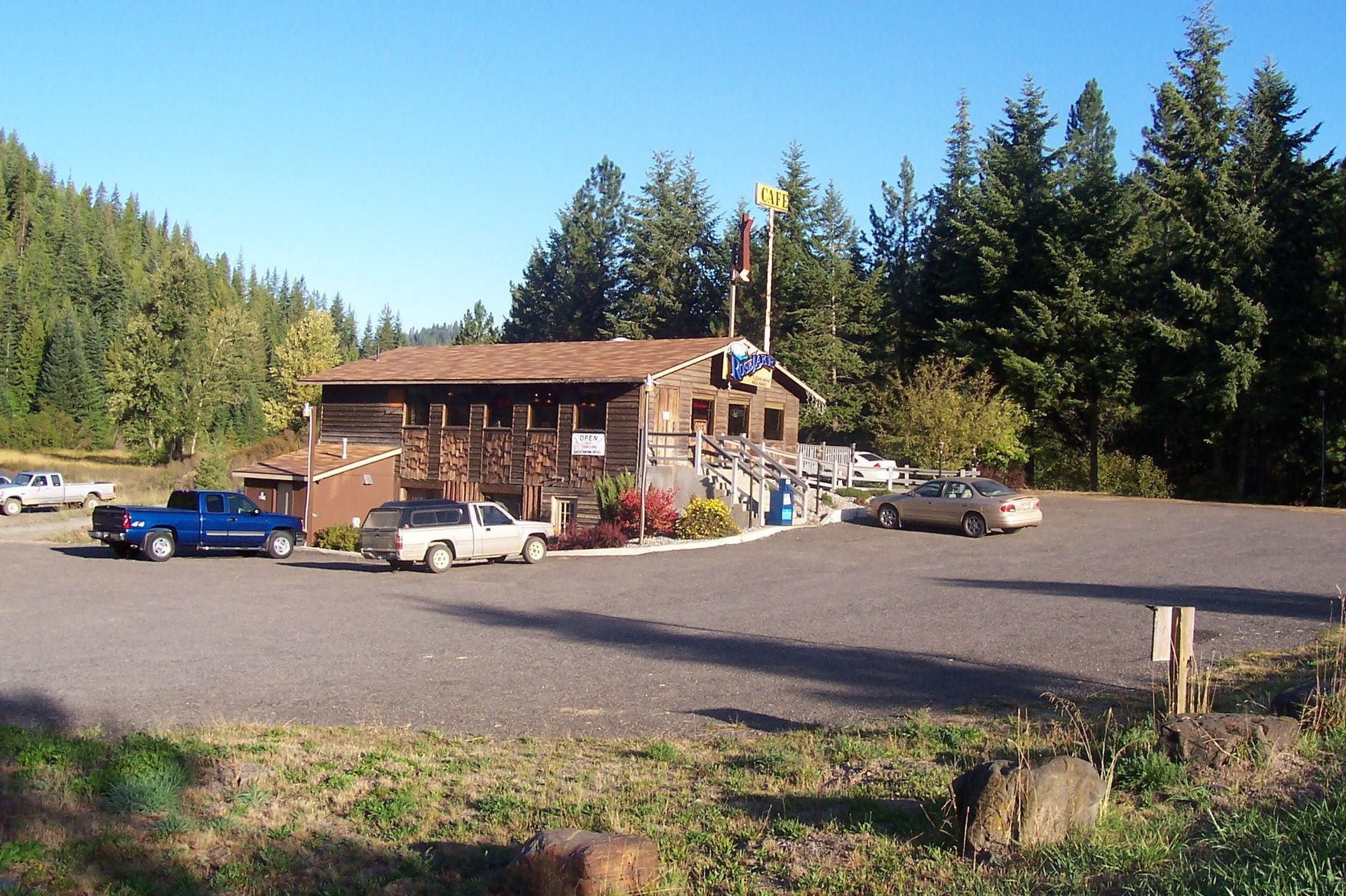 Rustic building with vehicles parked in front, trees in background, sunny day.