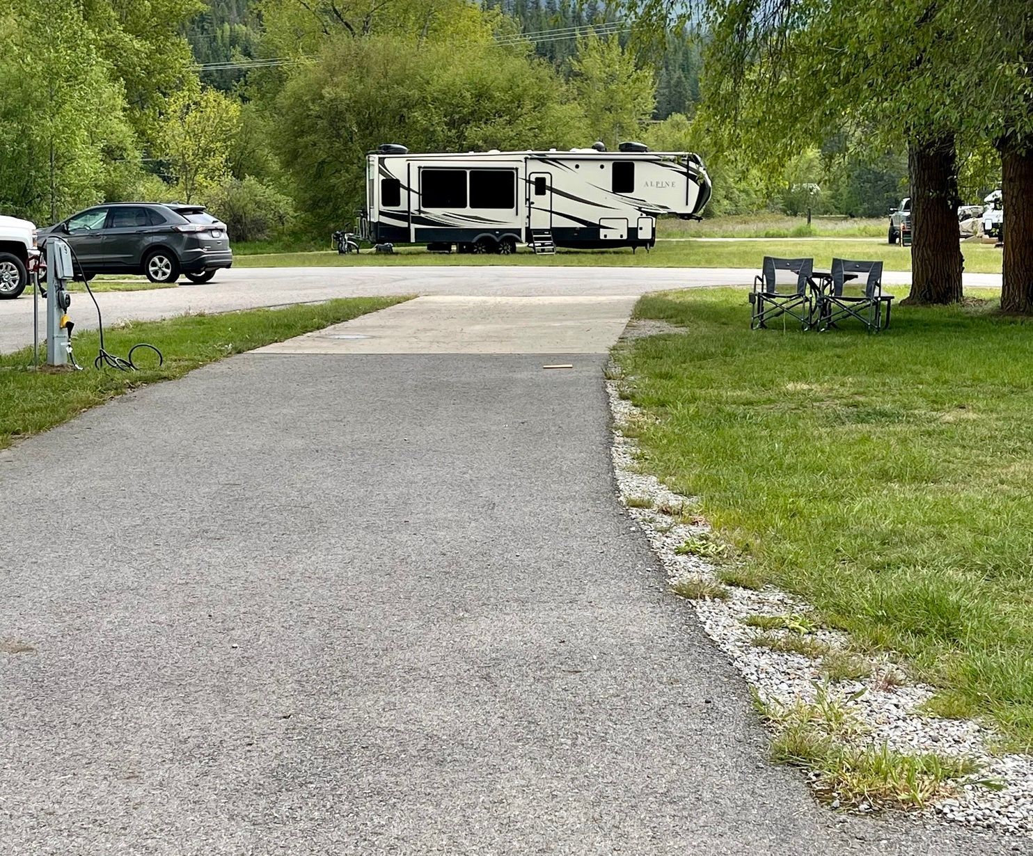 Gravel RV campsite with an RV, chairs, and a parked SUV near trees.