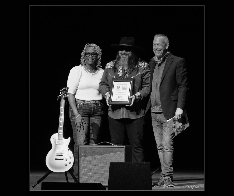 Three people onstage hold an award plaque beside a white electric guitar and an amplifier.