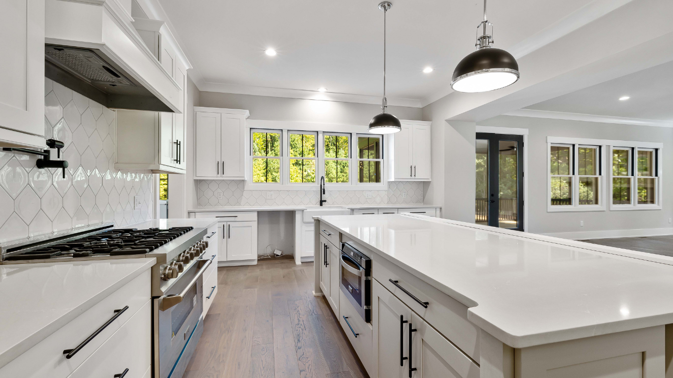 A bright, modern kitchen features a long white island, white cabinetry, a gas range, and hardwood floors.