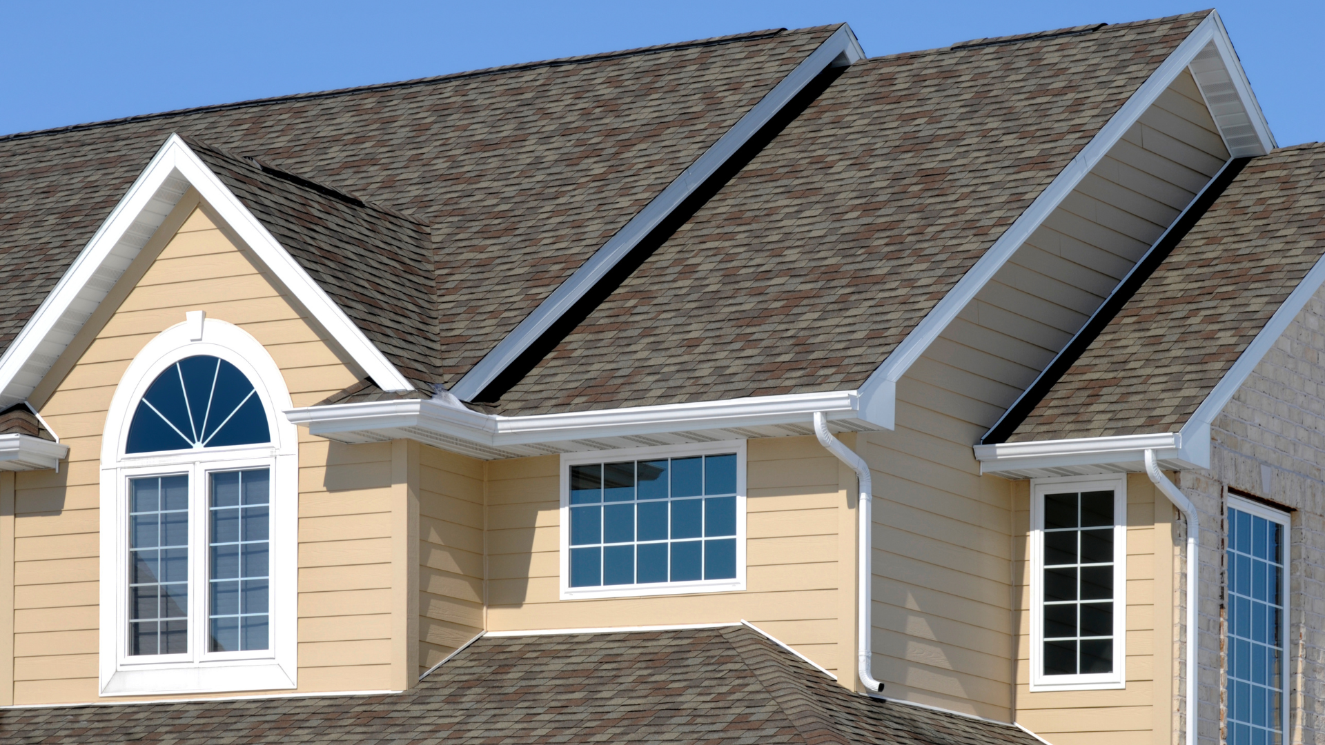 A tan, multi-story house exterior with brown shingles, white trim, and multiple rectangular and arched windows.