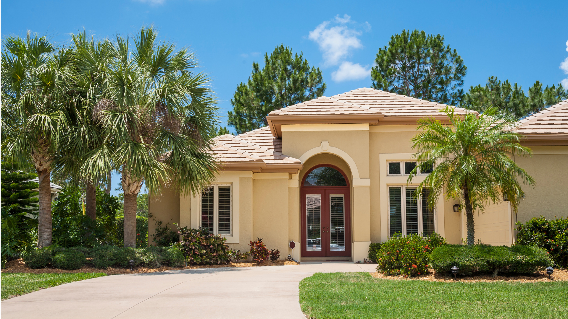 A single-story tan stucco house with a tiled roof, arched double front doors, and surrounding palm trees and green lawn.