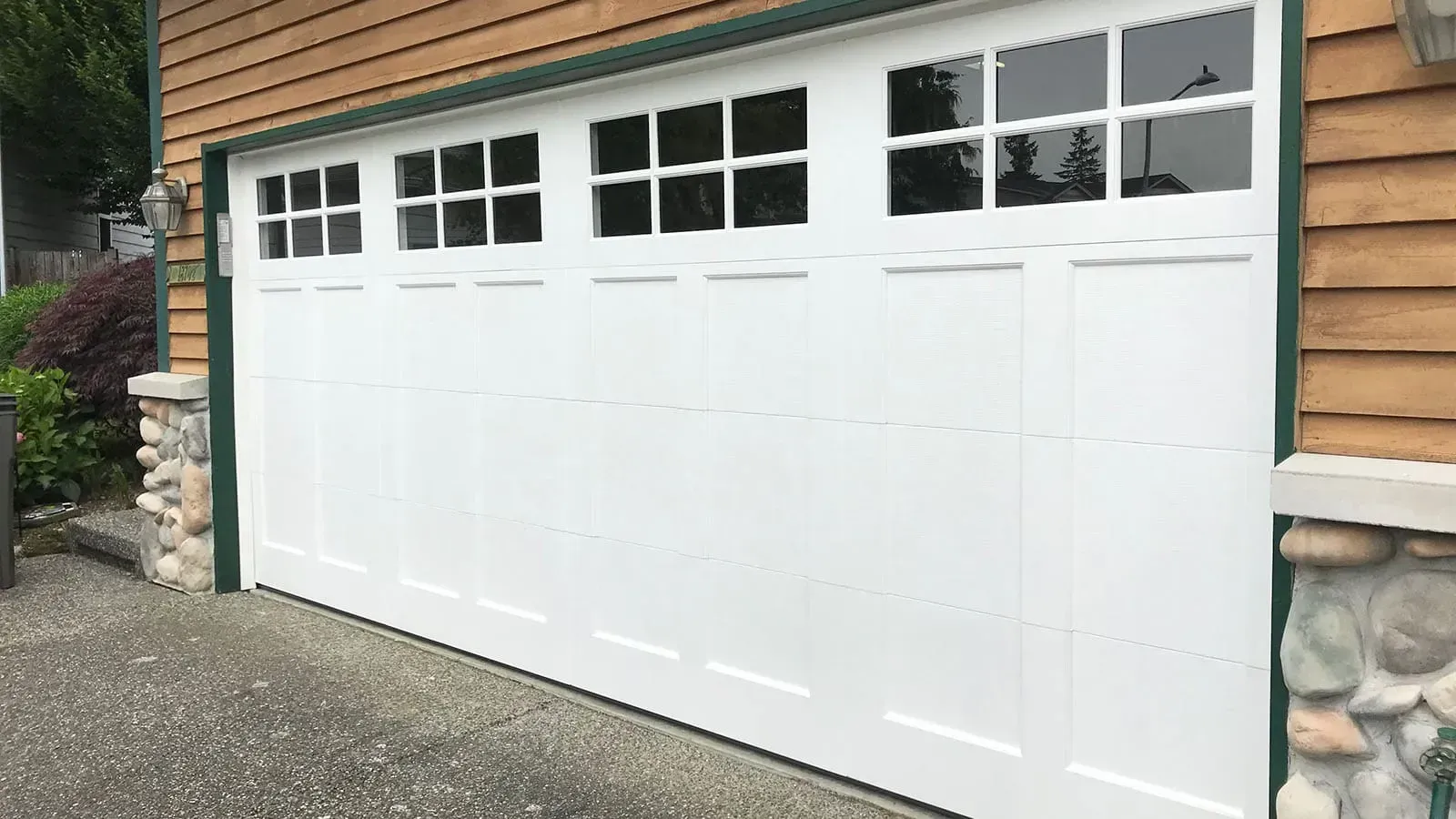 A white, paneled garage door with top window sections, mounted in a tan wood-sided home with stone corner accents.