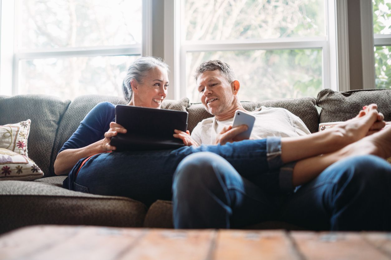 A couple relaxing on a couch, looking at a tablet and smartphone in a brightly lit room.