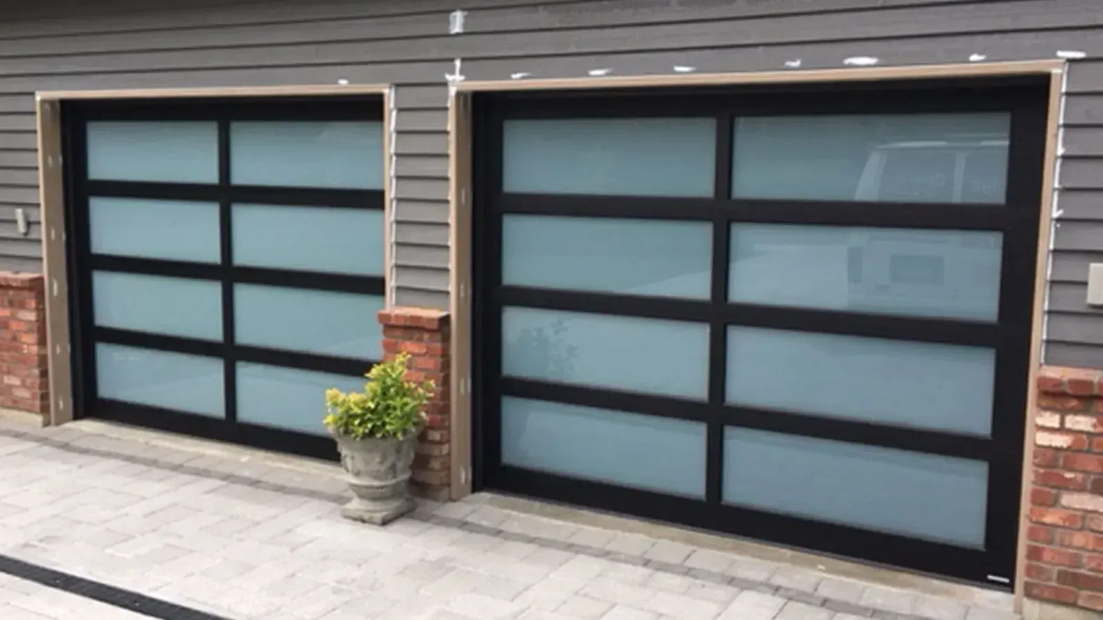 Two modern garage doors with black frames and frosted glass panels set in a grey house with a stone-accented driveway.