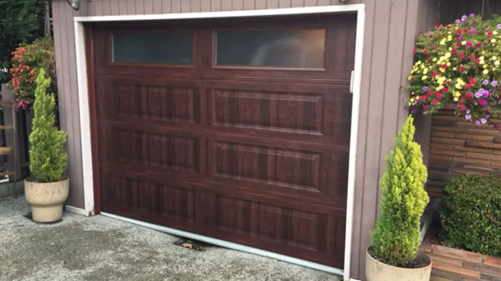 A dark brown, wood-grain garage door with two rectangular windows, flanked by two potted cone-shaped evergreen trees.