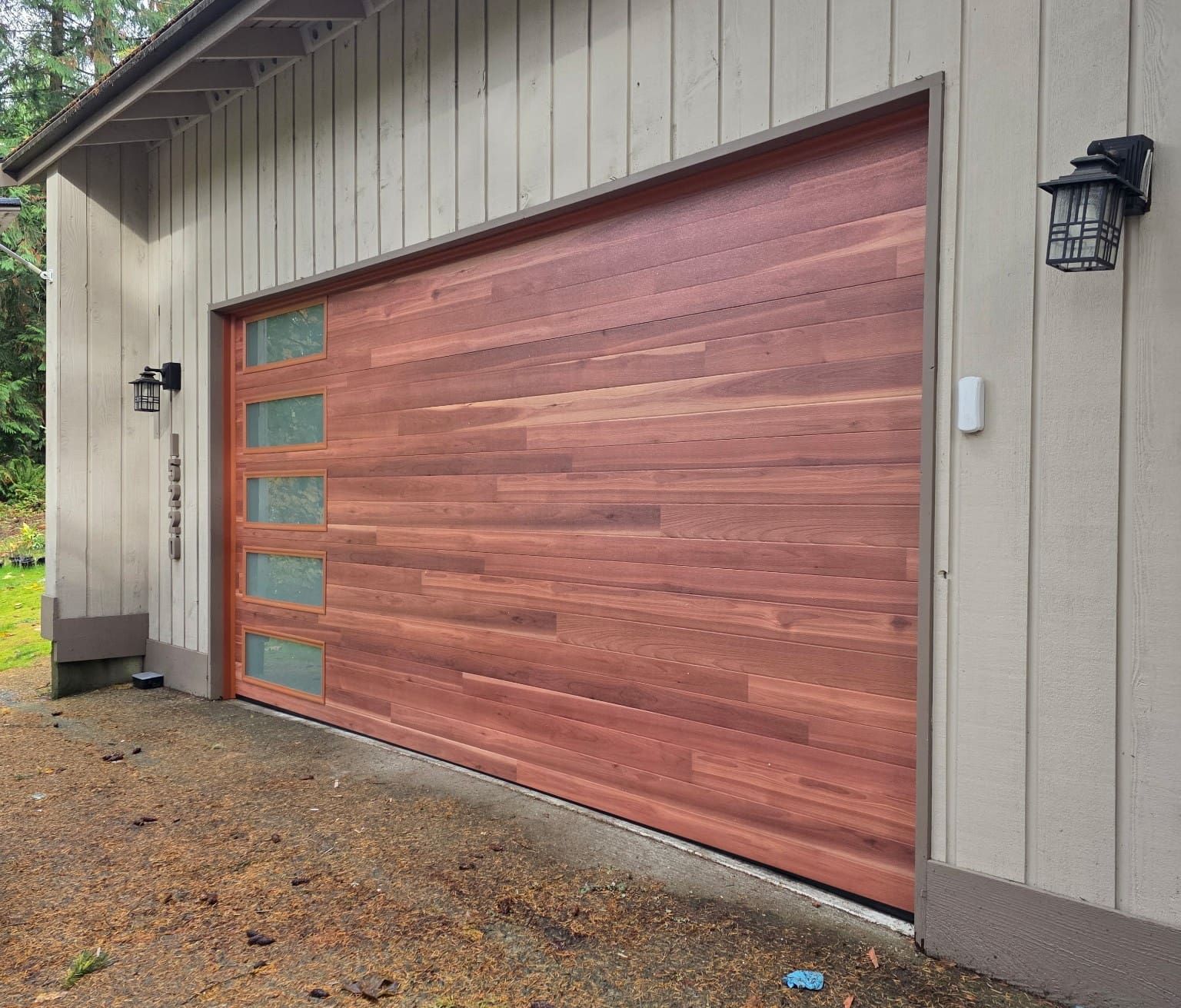 A wood-paneled garage door with a vertical strip of five rectangular frosted windows, set on a home with beige siding.