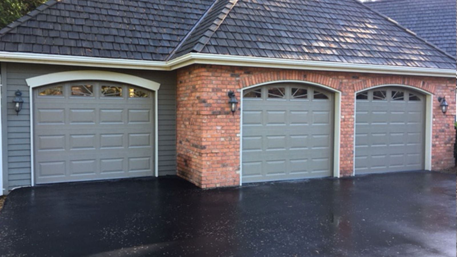 Three taupe-colored garage doors with arched windows set in a brick and gray-sided home with a dark shingled roof.