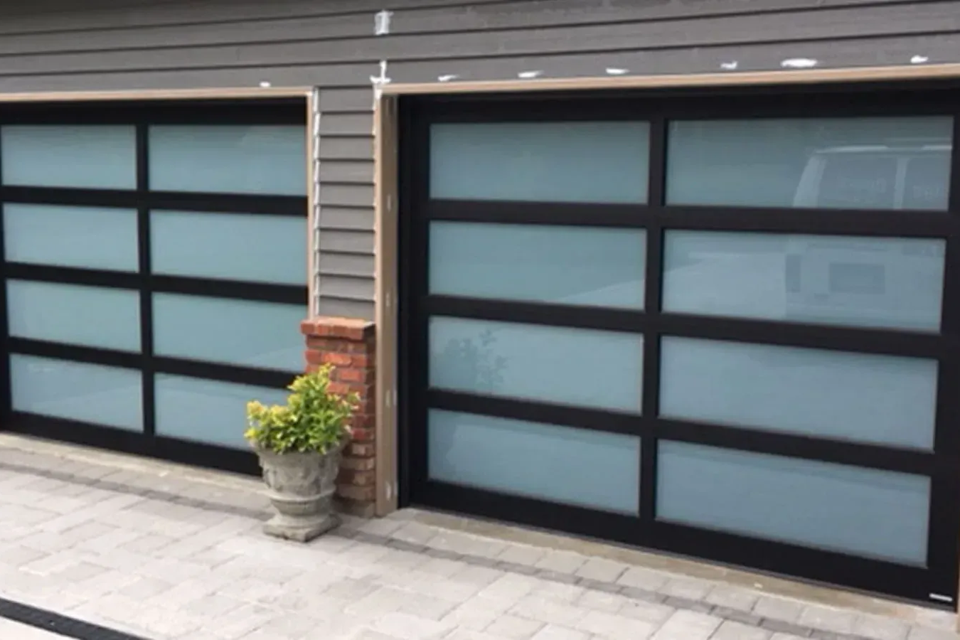 Two modern garage doors with black frames and frosted glass panels, set against gray horizontal siding.