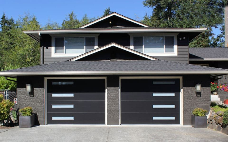 A two-story house with dark gray brick, two modern garage doors with horizontal windows, and surrounding trees.