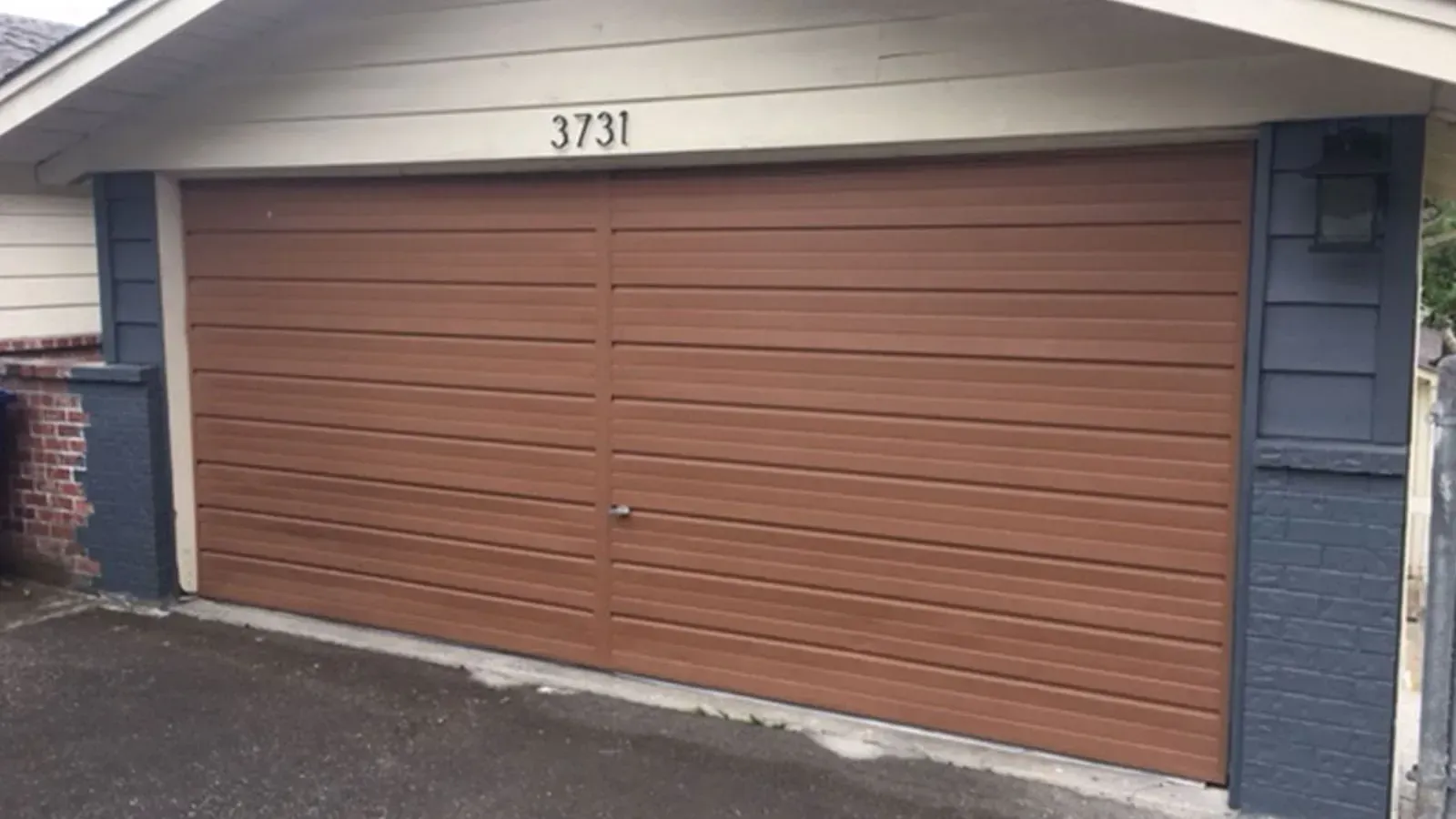 A brown, horizontal-paneled garage door with the number 3731 above it, flanked by dark grey siding and stone pillars.