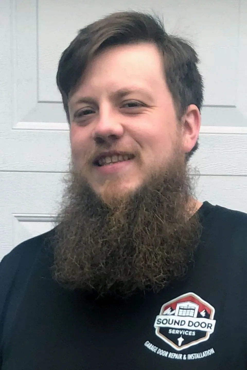 A Sound Door Services technician with a full beard wearing a black work shirt with a garage door service logo.