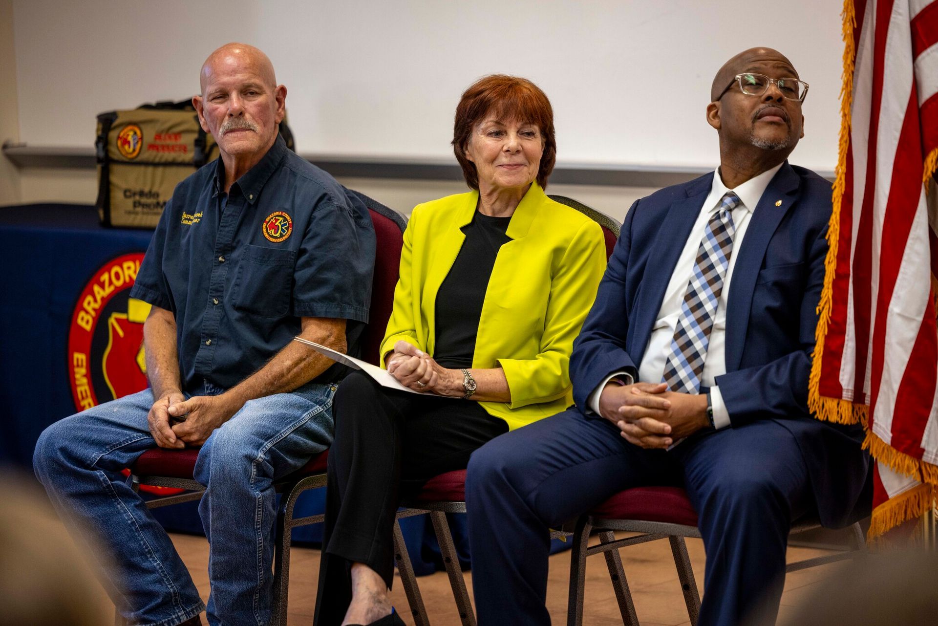Three people are sitting in chairs in front of an American flag at an announcement meeting.