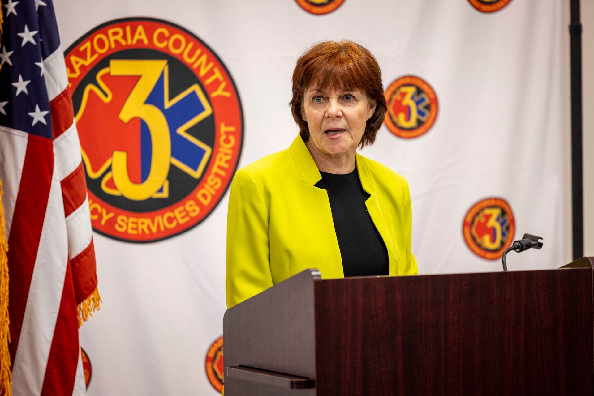 A woman in a yellow jacket is standing at a podium in front of an American flag.