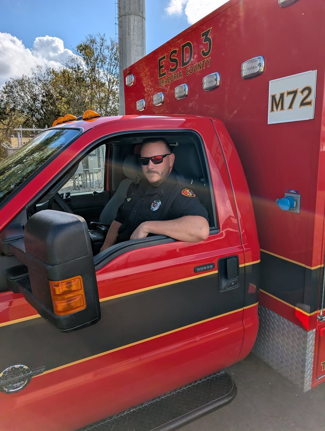 Red emergency vehicle with a driver in the cab, parked outdoors near an M72 sign.