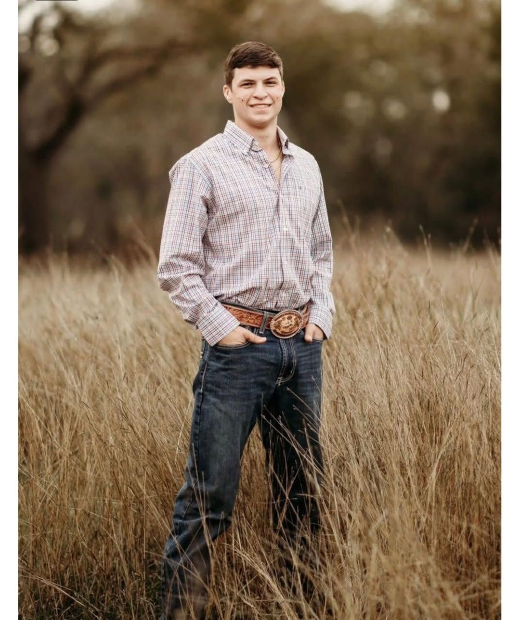 Austin Stallman standing in tall grass outdoors, wearing a patterned shirt, jeans, and a large belt buckle, smiling at the camera