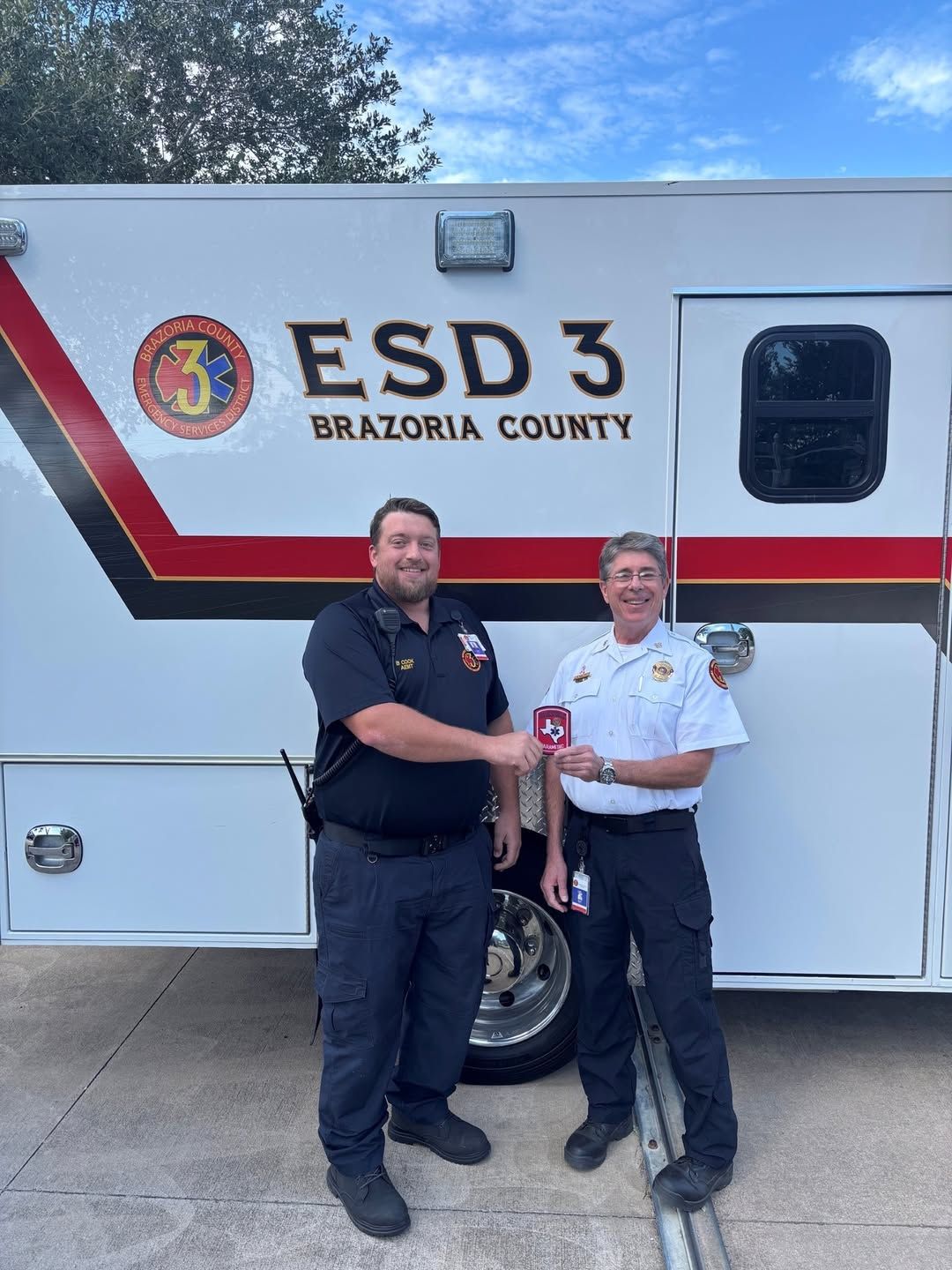A paramedic and EMS Chief standing beside a white emergency vehicle, shaking hands and smiling.