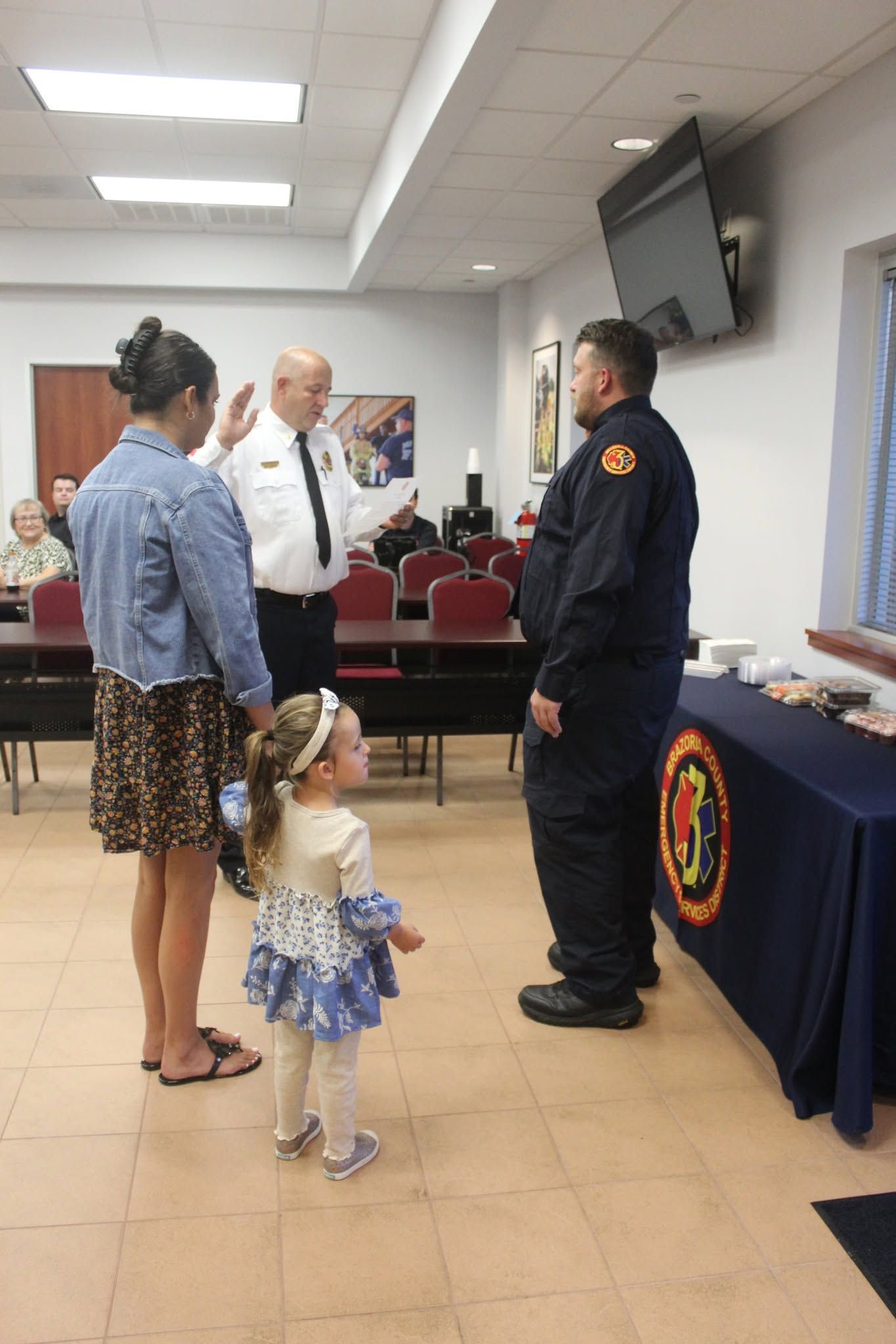People standing at a ceremony in a hall, with a uniformed officer beside a table with a department emblem.