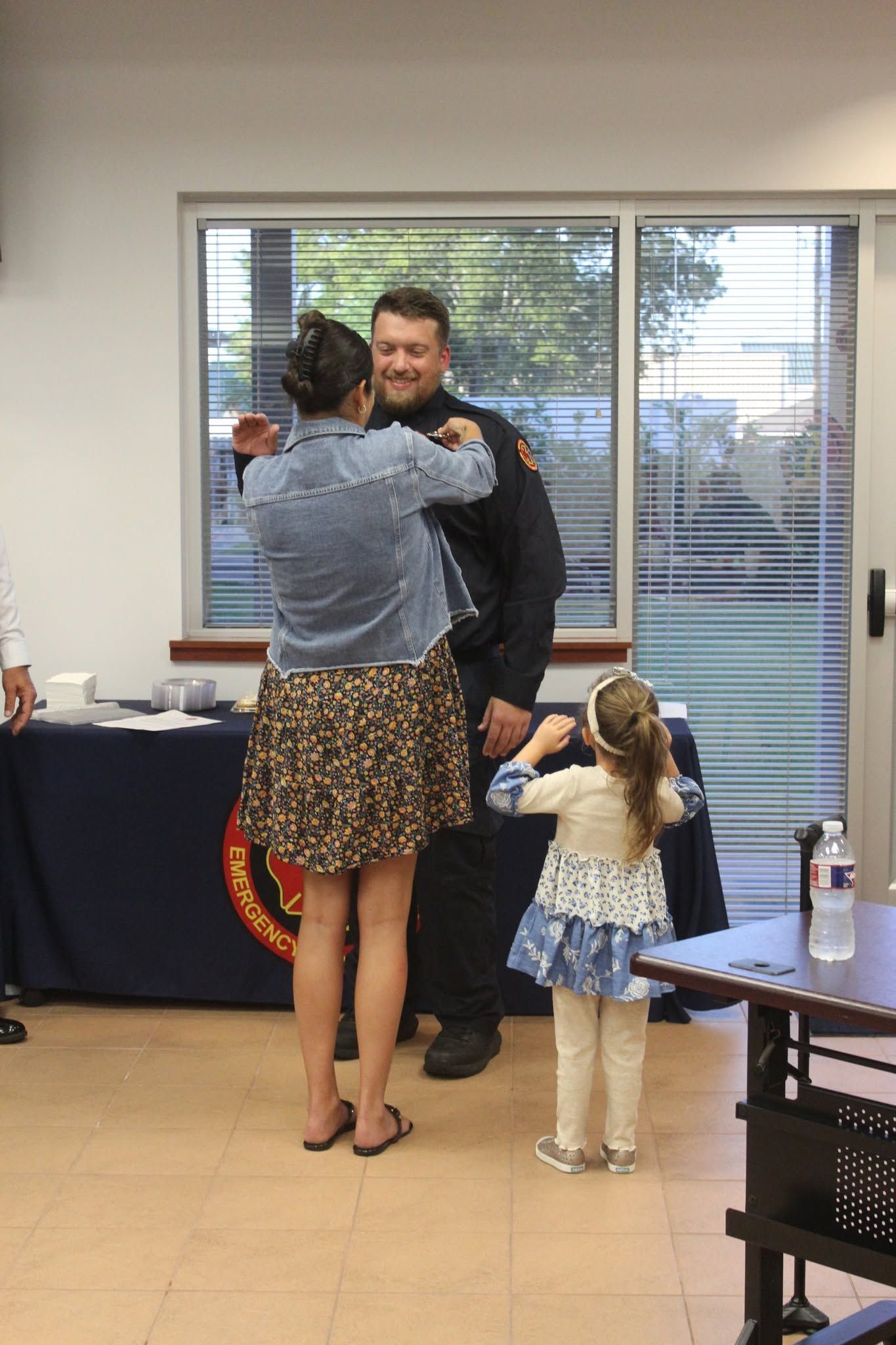Two people hugging in an office while a small child stands nearby by a table and large windows.