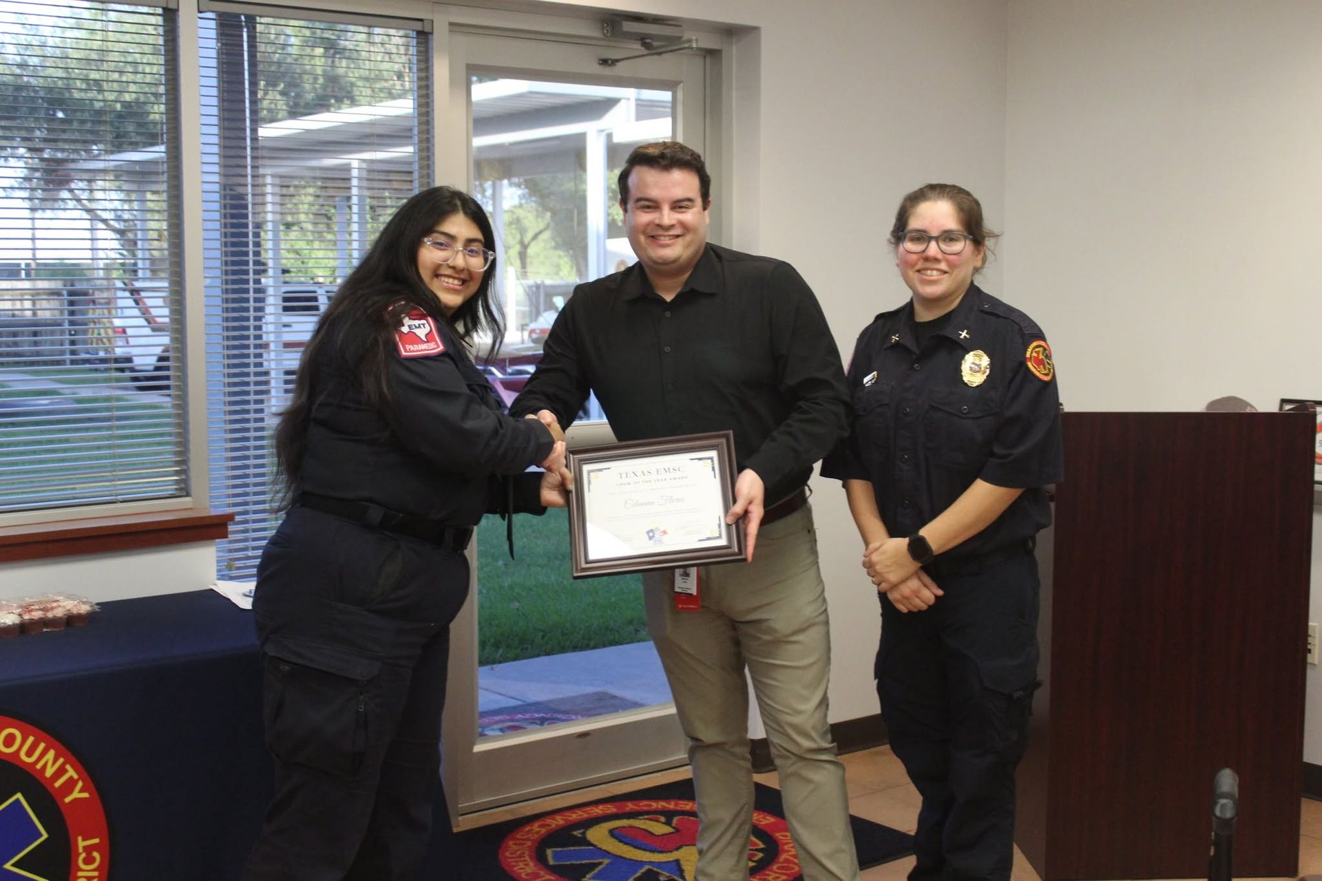 Three people in a room hold a framed certificate, smiling beside a table and doorway.