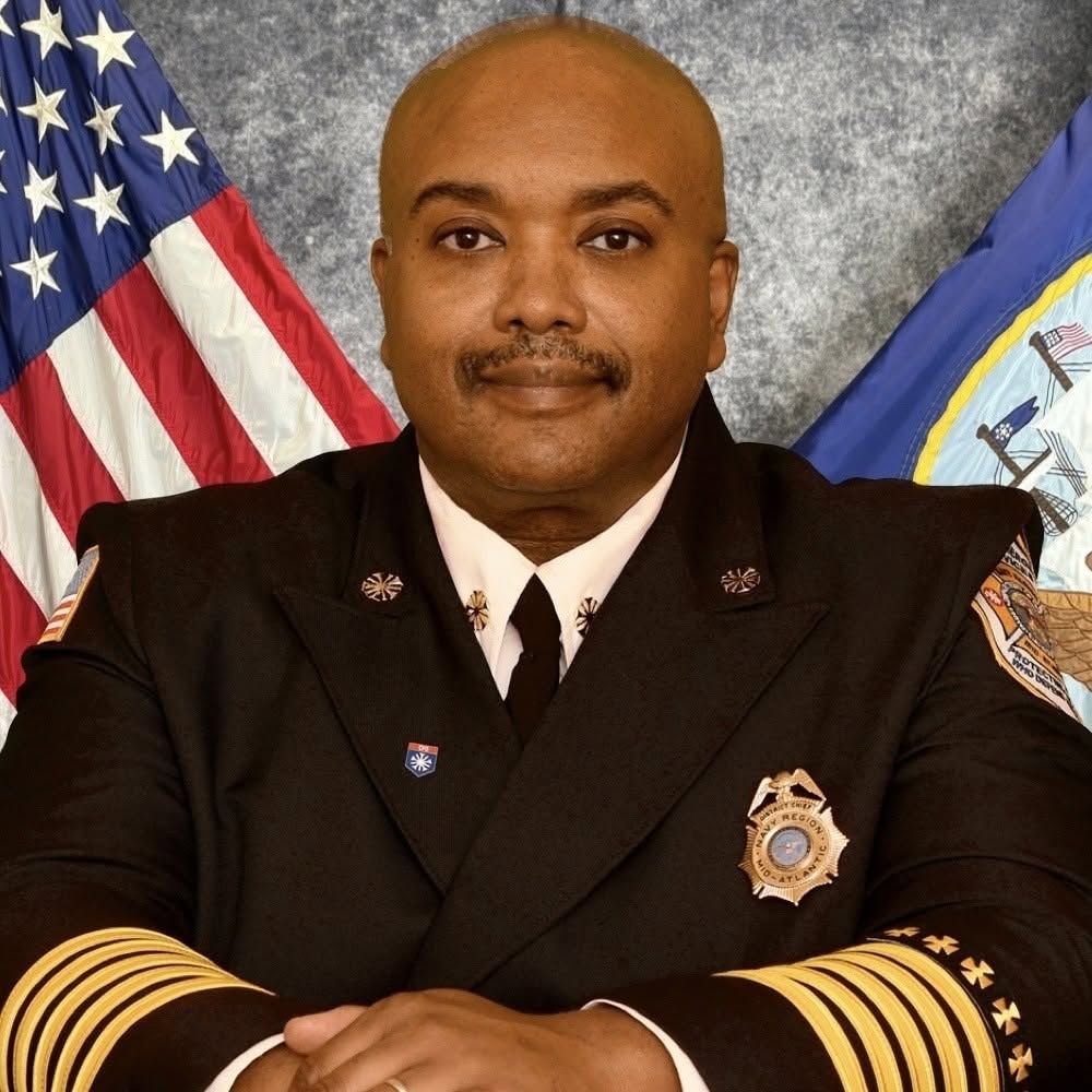 Uniformed officer posing with arms crossed in front of American and state flags