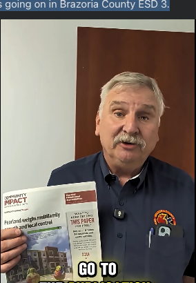 Man holding an informational brochure and speaking indoors, wearing a dark shirt with a logo badge.