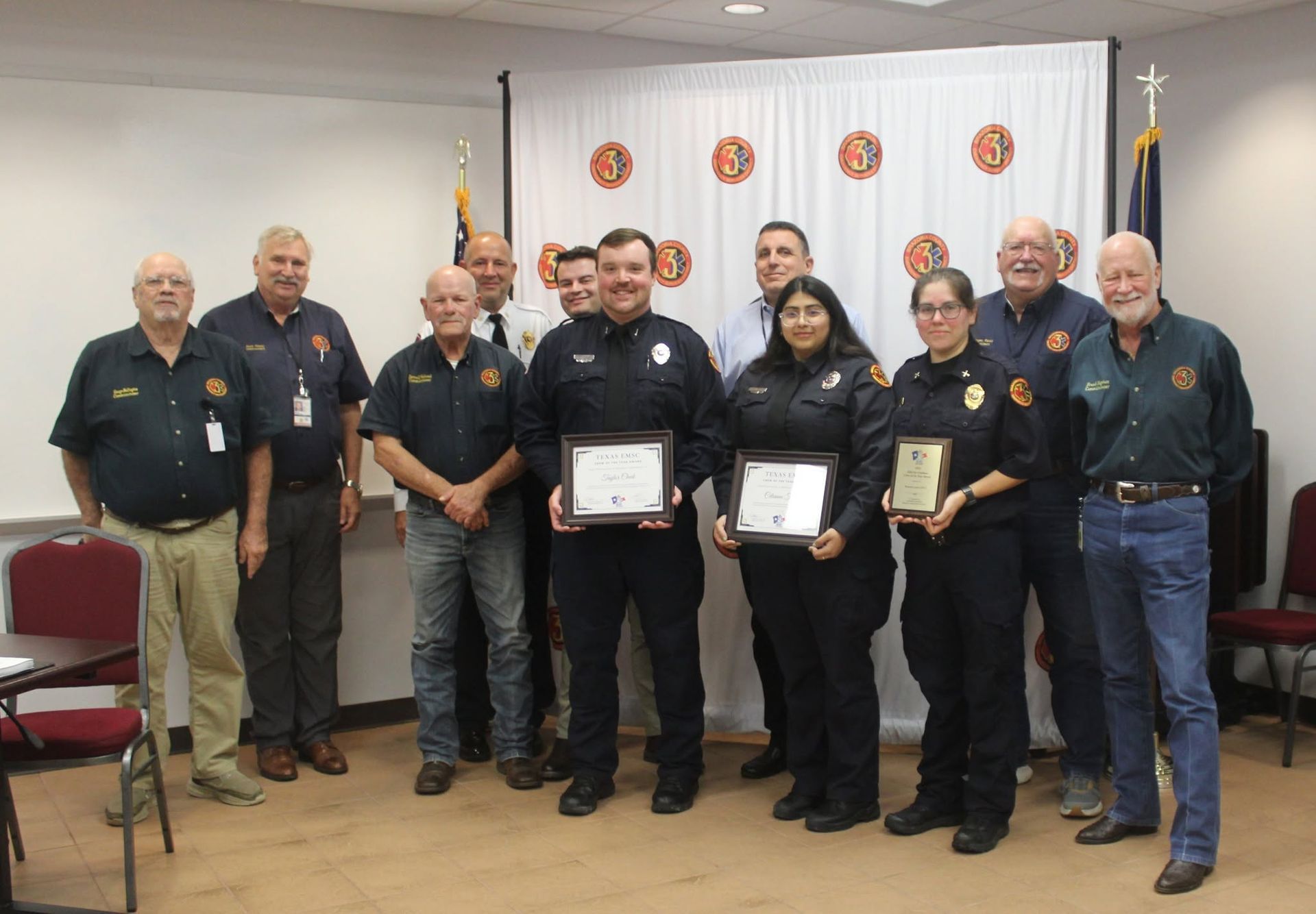 Group of uniformed firefighters posing indoors, three holding framed awards in a ceremony room