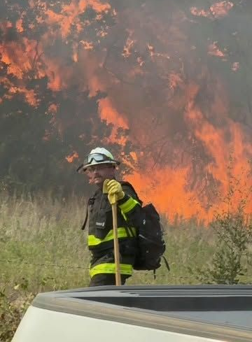 Firefighter in yellow gear stands near tall flames and smoke in a grassy field.