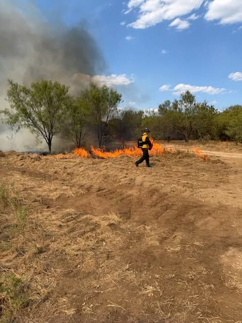 Firefighter battling a grass fire in a dry field, with smoke rising under a blue sky.