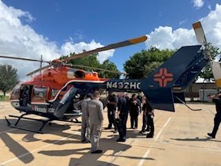 People gathered beside an orange-and-blue helicopter on a sunny tarmac