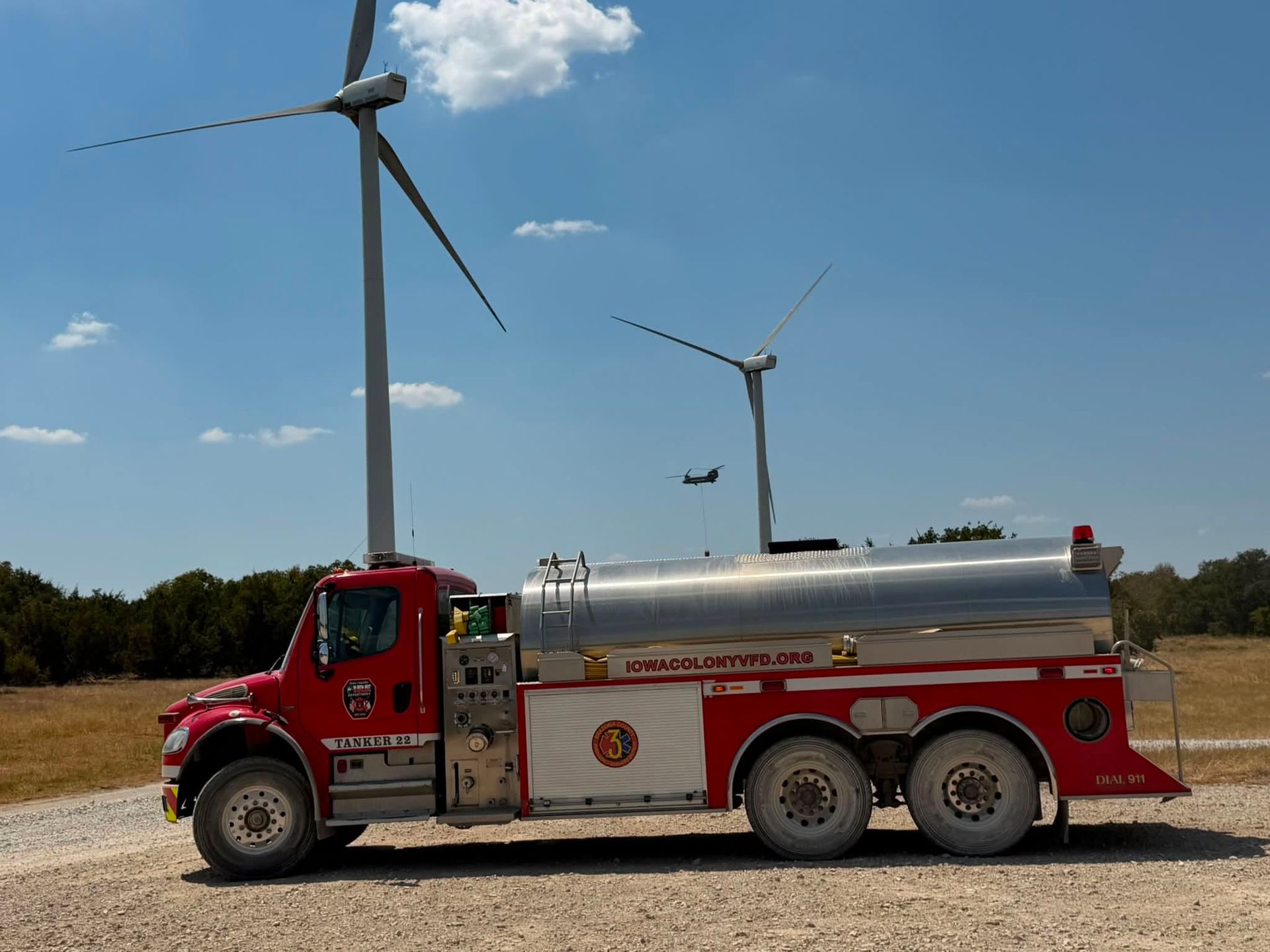 Red Iowa Colony tanker truck parked on a dirt road near wind turbines under a blue sky.