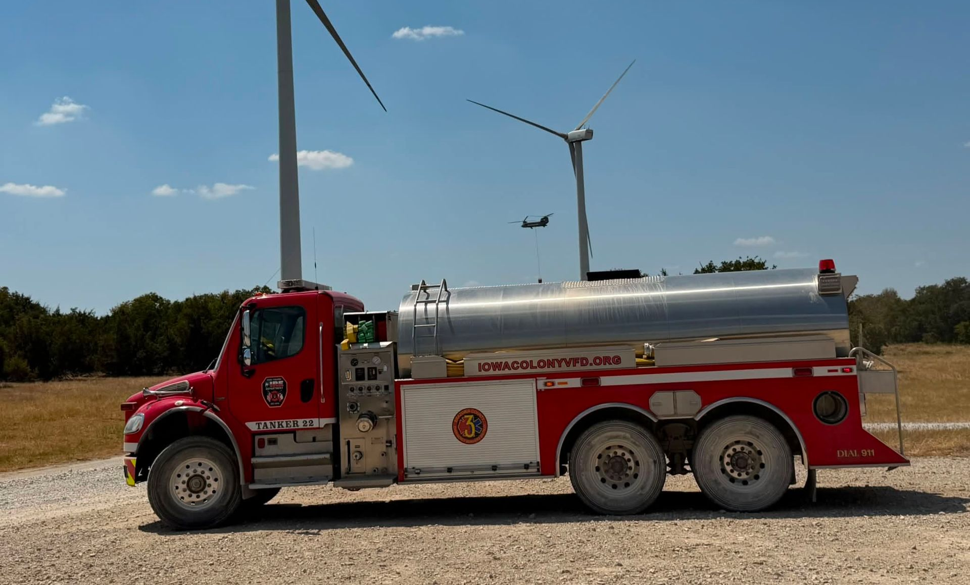 Red Iowa Colony fire tanker truck parked on dirt beside wind turbines under a clear blue sky