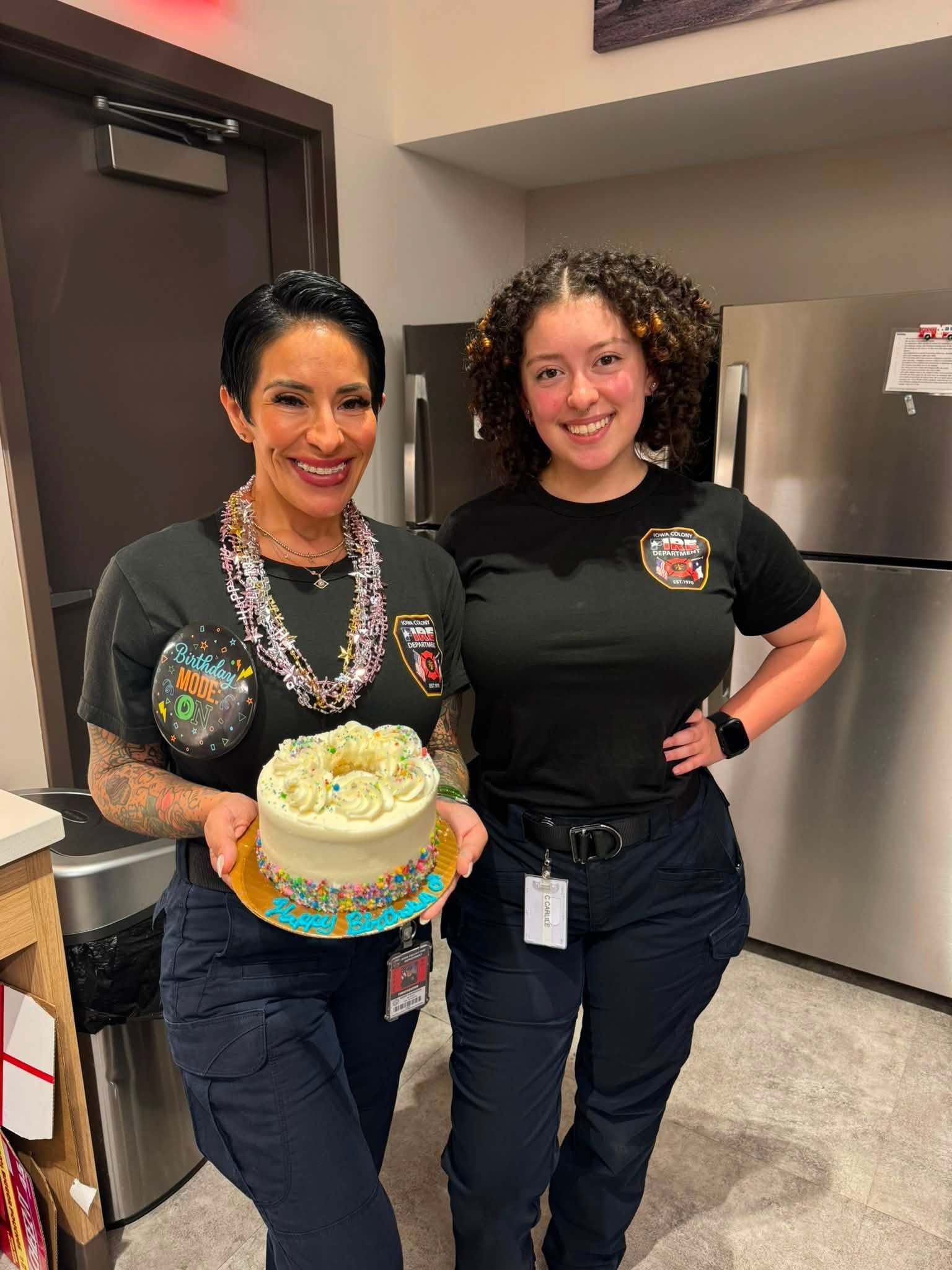Two smiling staff members hold a decorated cake in a kitchen break room.