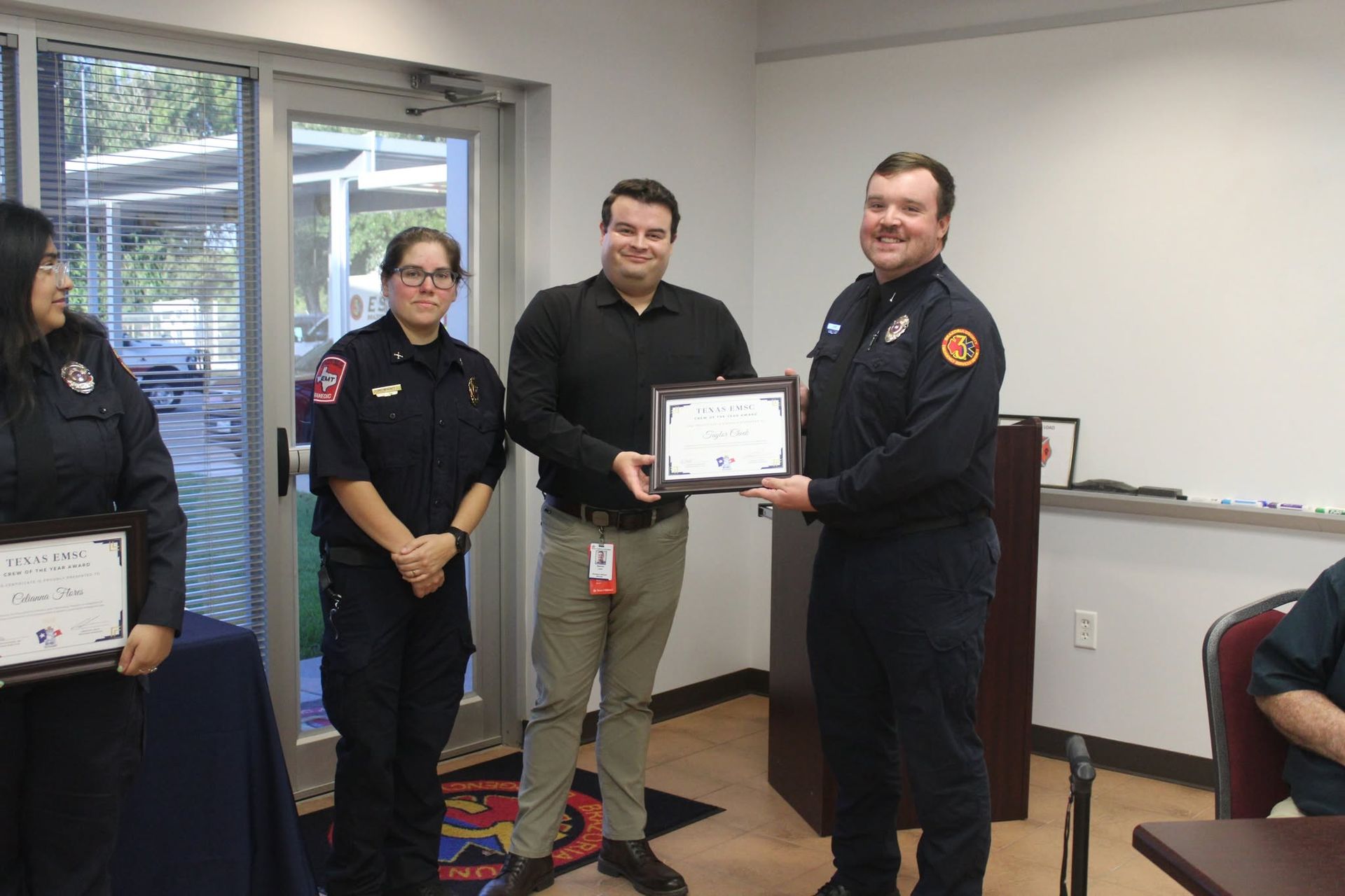 People in uniforms and casual clothes stand in an office holding framed certificates.