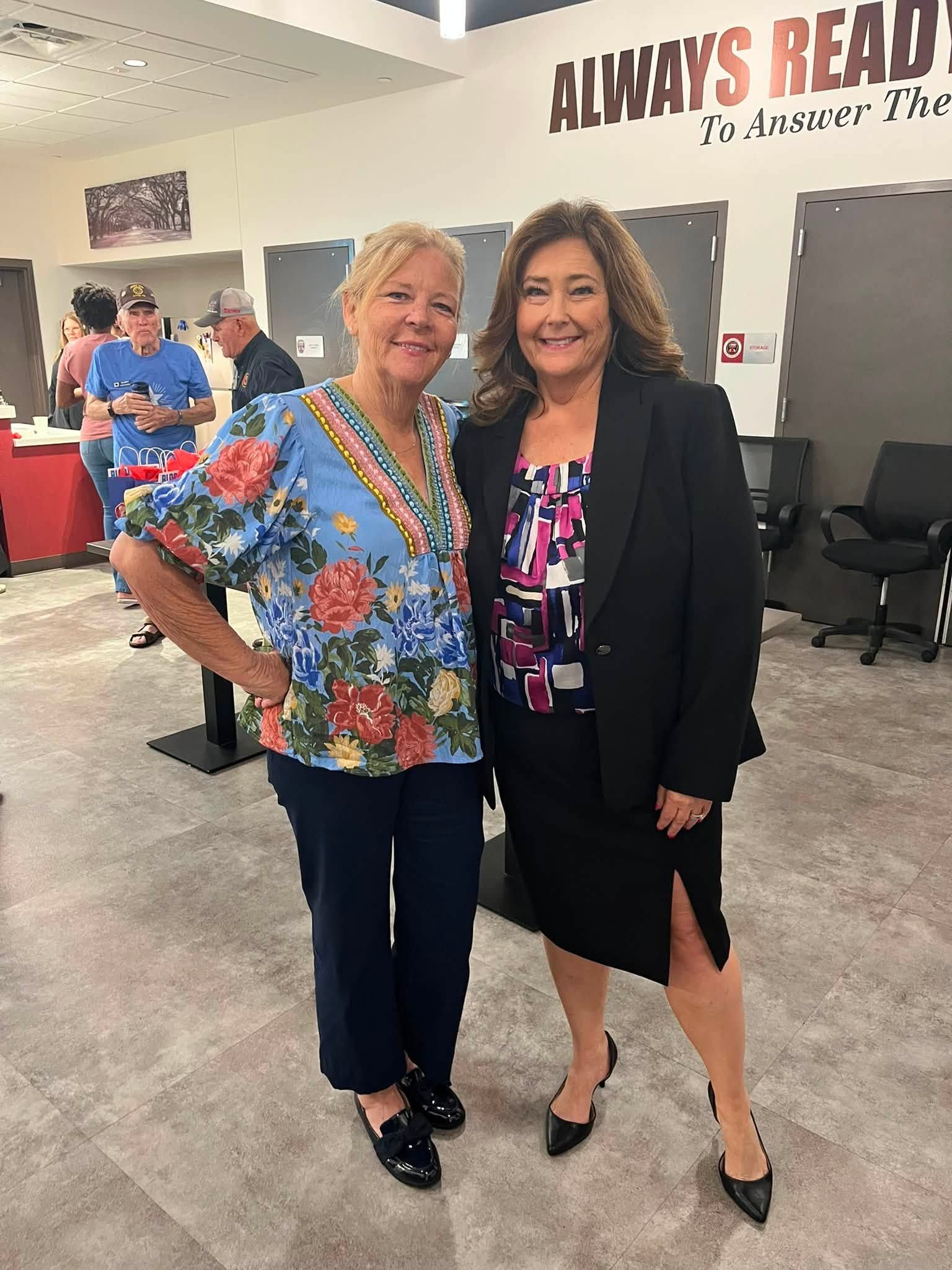 Two women pose smiling in an office lobby, one in a floral blouse and one in a black blazer.