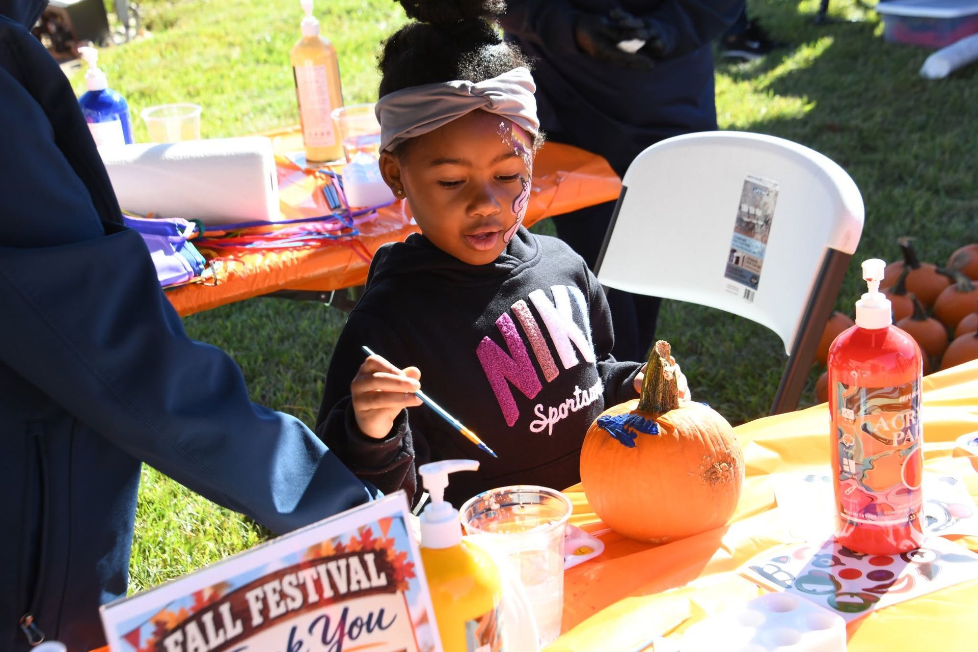 Child at a fall festival painting a pumpkin at an orange table, wearing a black shirt.