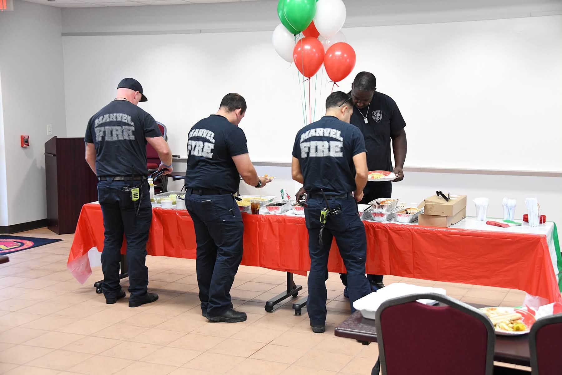 Three firefighters stand at a red-covered table with supplies and balloons in a room.
