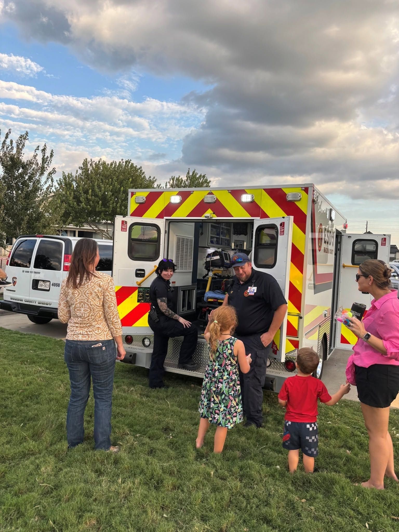 People gathered around an open ambulance with red-and-yellow chevrons in a grassy field.
