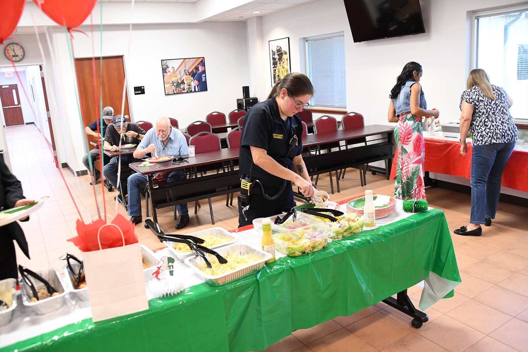 People serving food at a buffet table in a community room with balloons and chairs