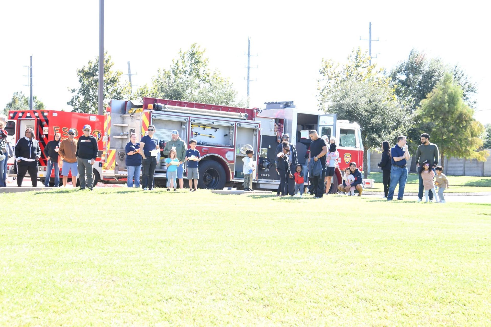 People gathered around a red fire truck on a grassy field in bright daylight.