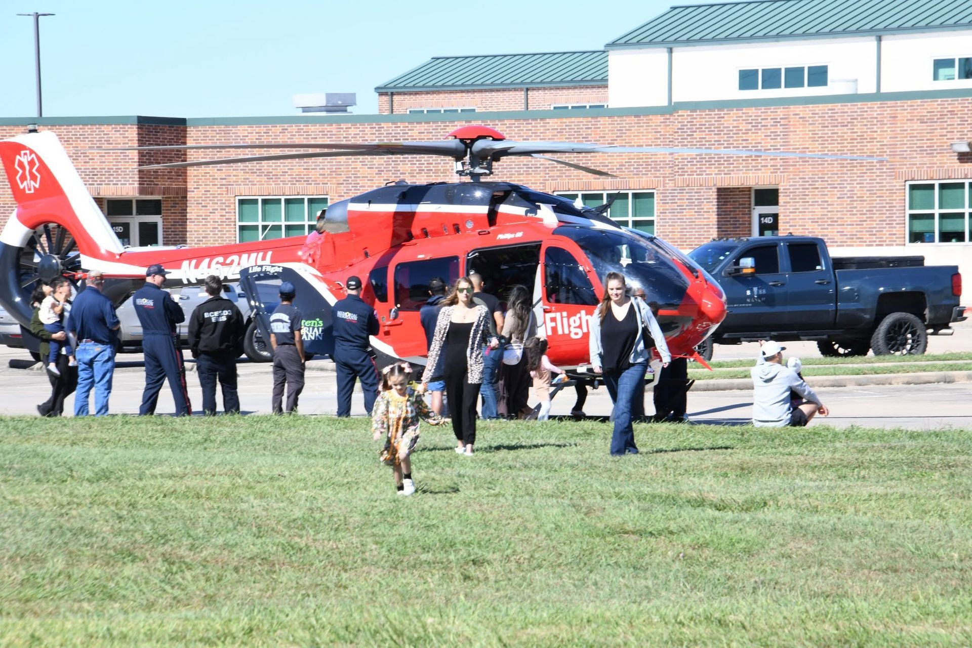 Red rescue helicopter on grass by a brick building, with firefighters.