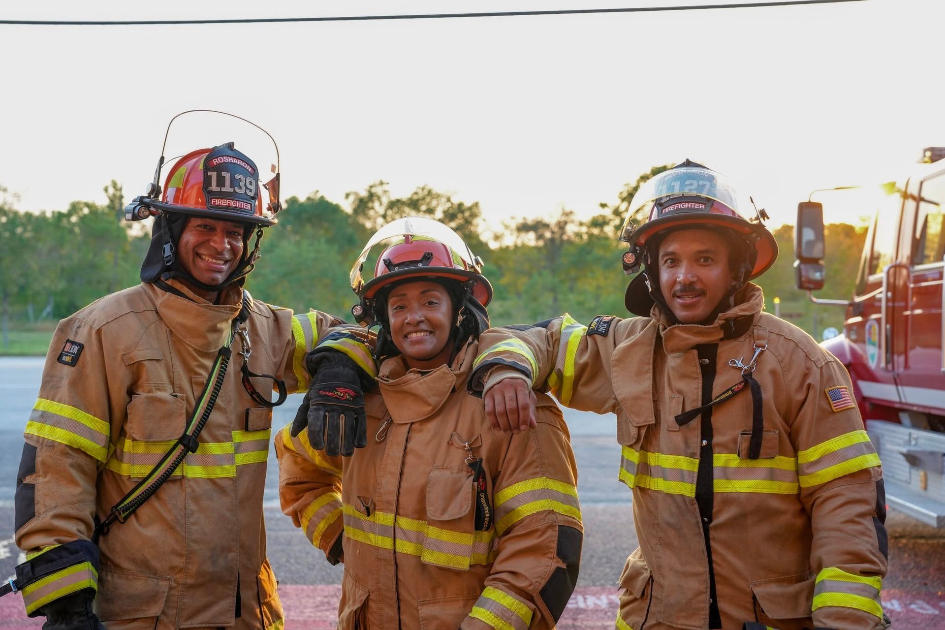 Three firefighters in tan turnout gear and helmets smiling outdoors at sunset