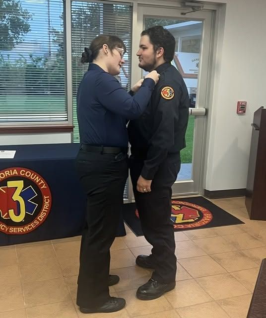 Two emergency responders standing face-to-face indoors, one adjusting the other's collar near a glass door.