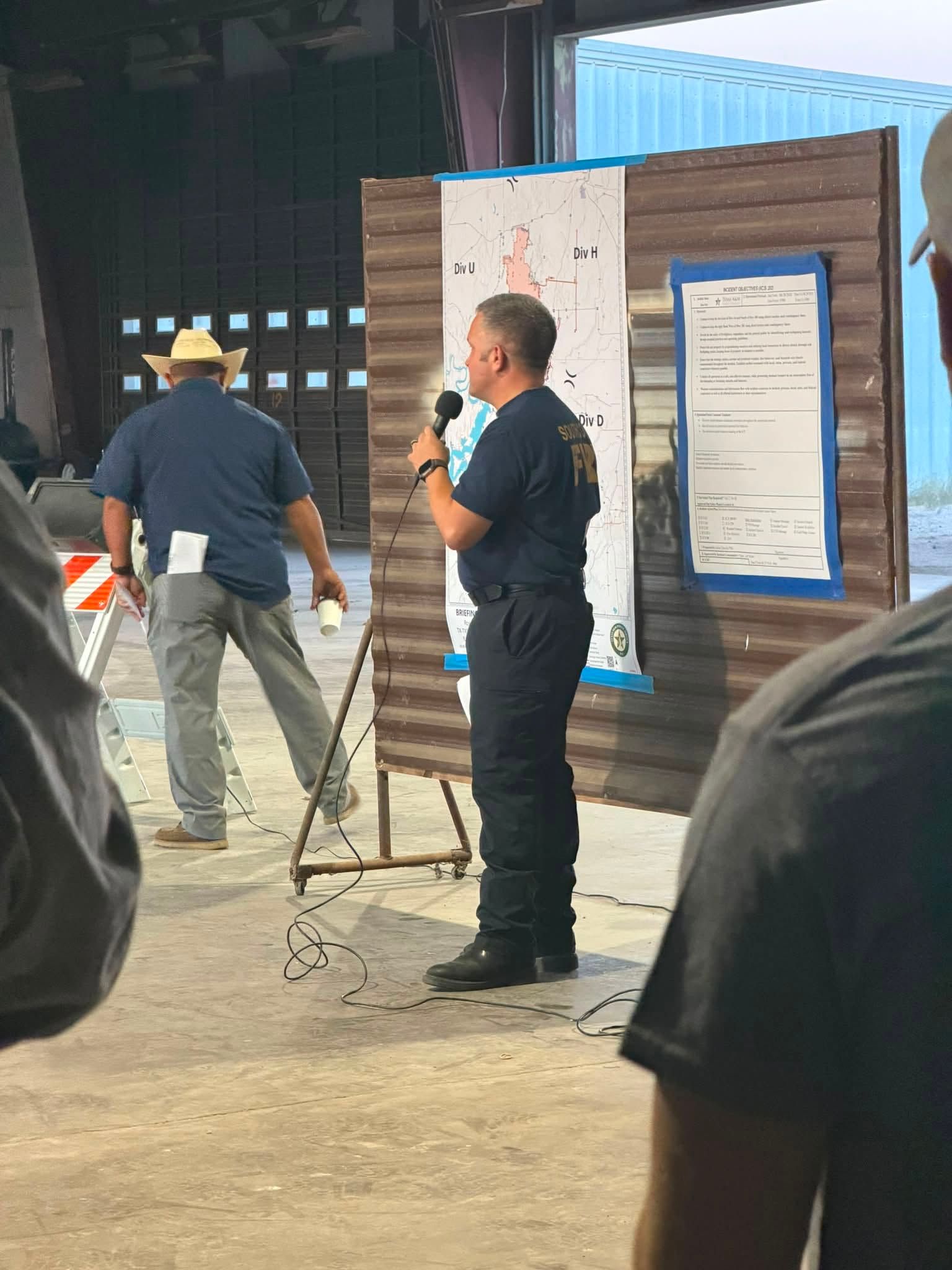 Two men speaking at a Ross Fire breifing, one holding a microphone beside a poster display on an easel.