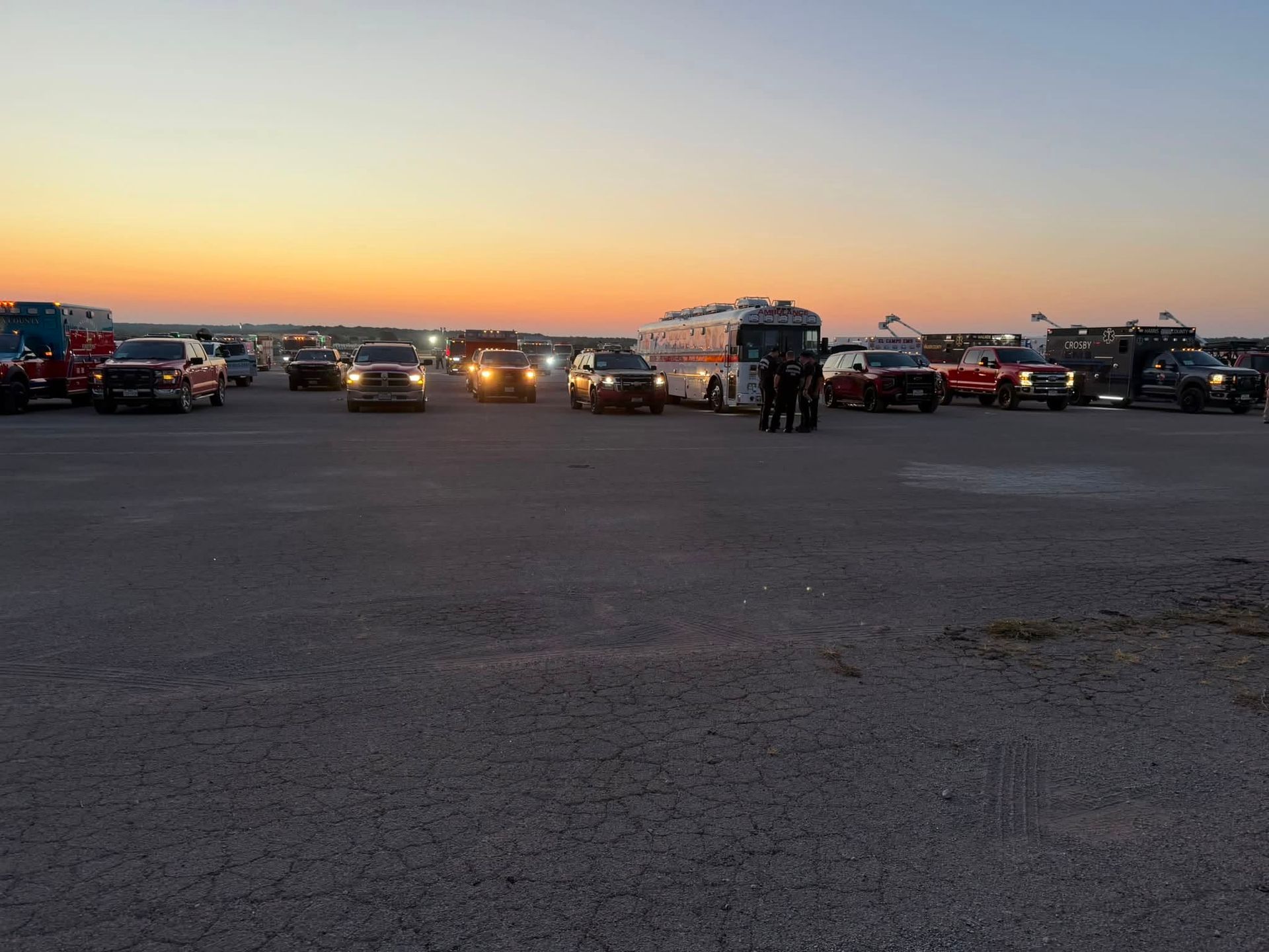 Cars parked in a lot at sunset, with headlights on and a few people standing near the center.